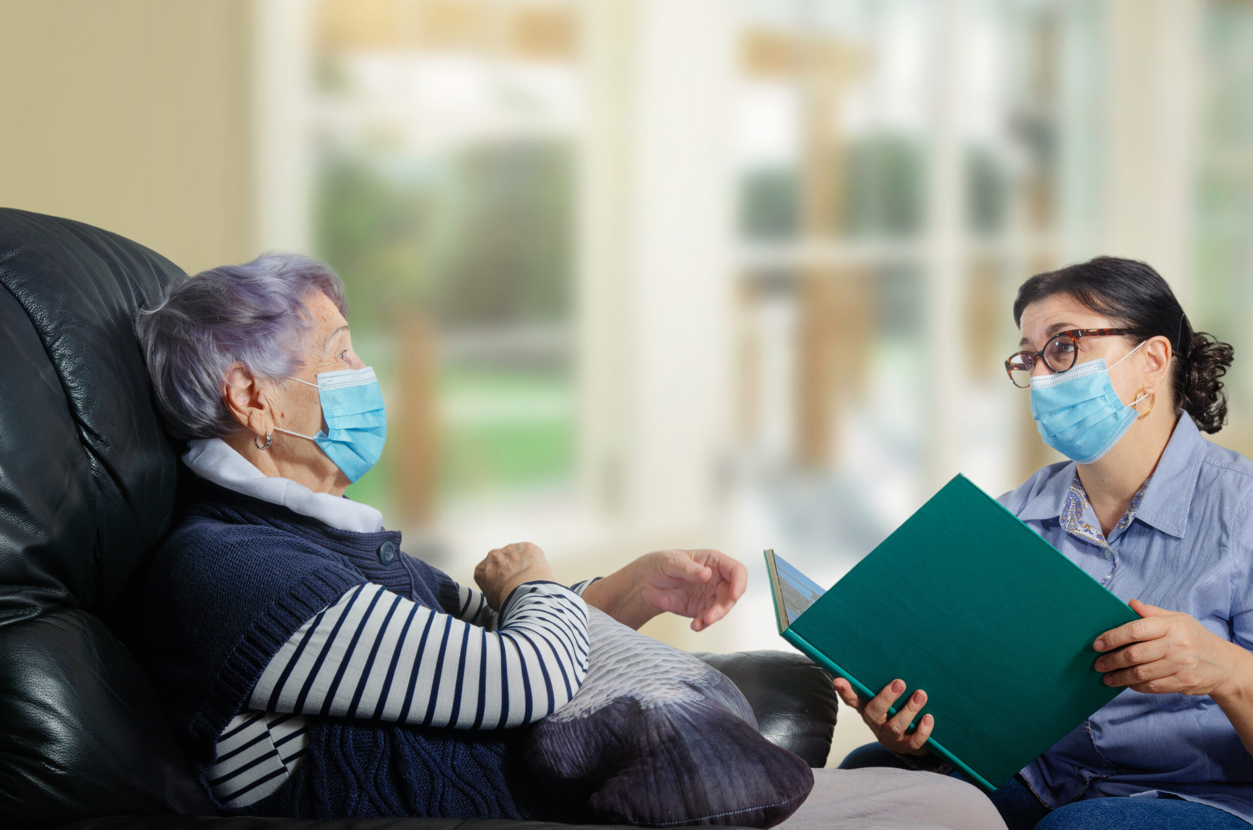 A female adult volunteer visits a single senior woman during a pandemic. She talks to her and reads books. Both wear face protective masks.