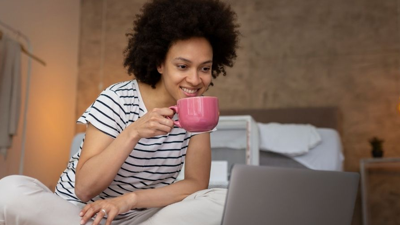 young woman sitting on the floor, looking at her laptop with a pink mug in her hand