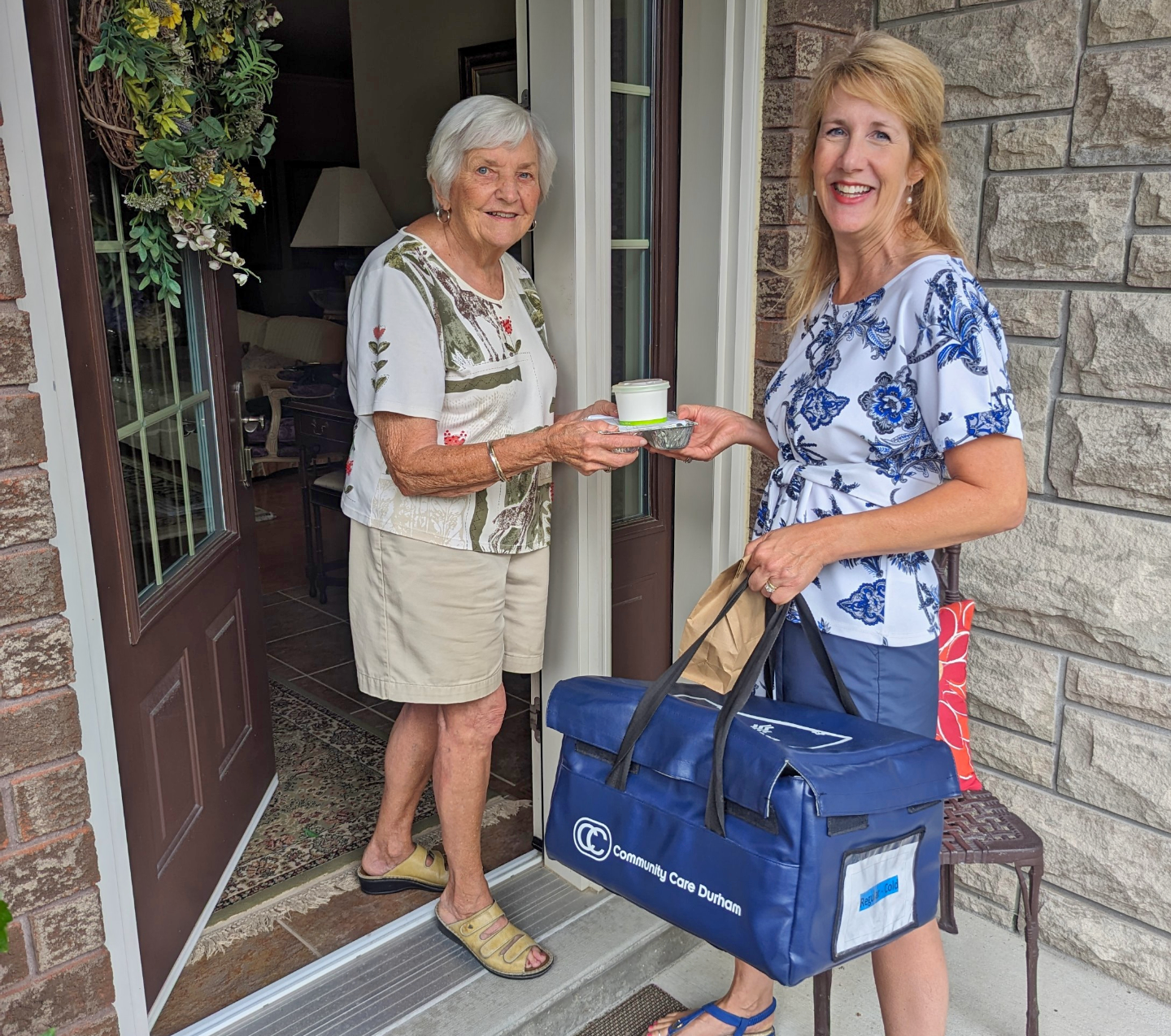 Client accepting Meals on Wheels deliver from volunteer. Both smiling for photo.