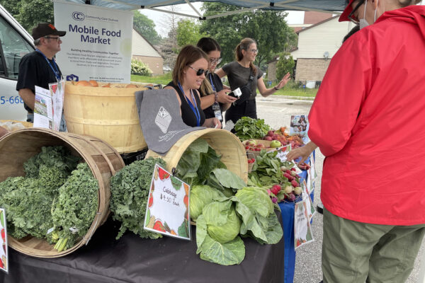 Photograph of Mobile Food Market team behind table taking payments from supporters. Table has fruits (apples, strawberries) and vegetables (lettuce, kale, onions) on display.