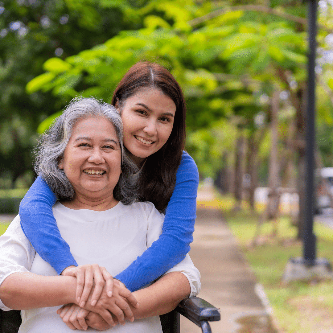 Photo of woman in blue shirt hugging woman in white shirt from behind outside on sidewalk. Mother is sitting in wheelchair.
